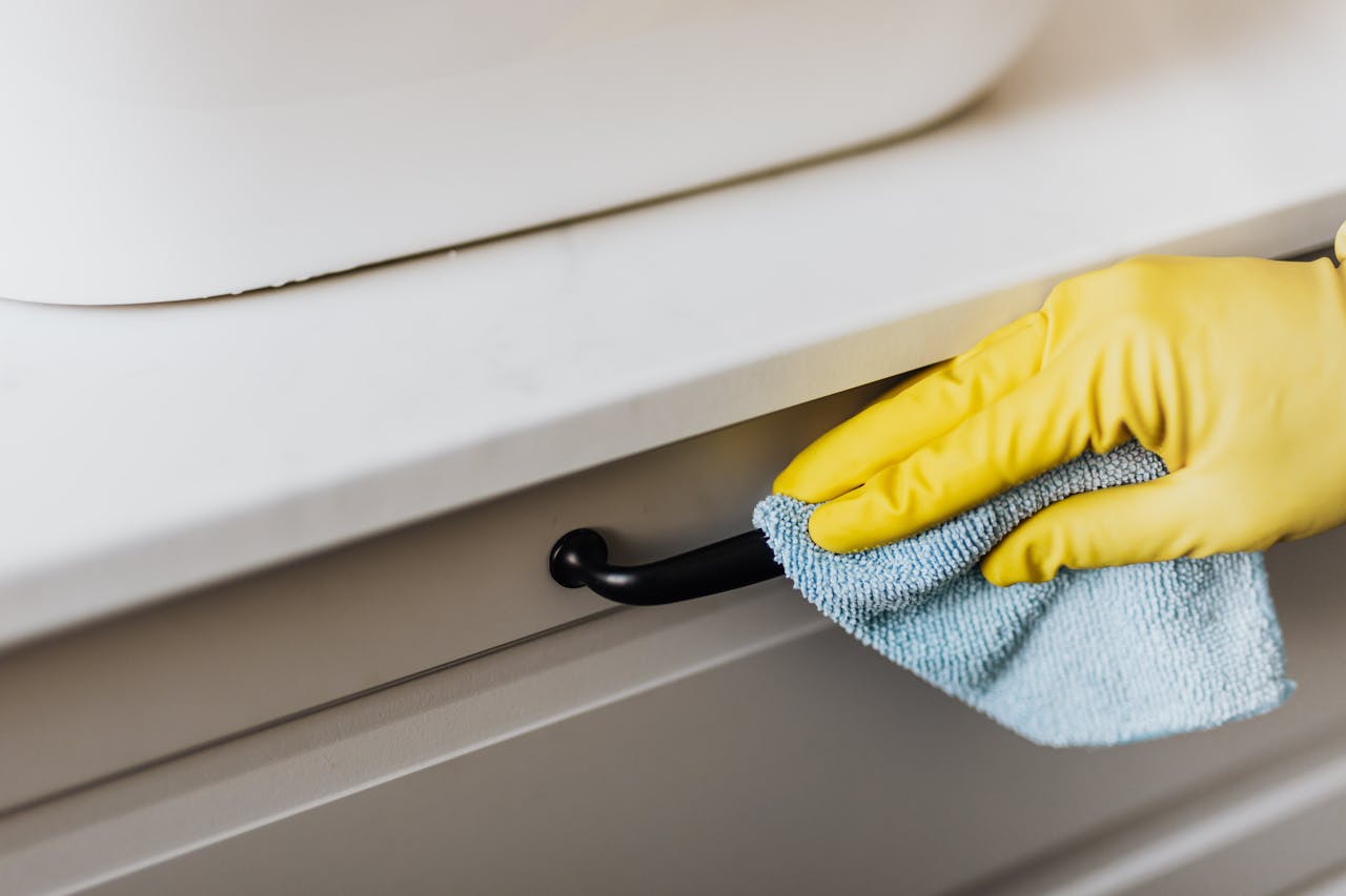 Contact From above of crop faceless person in yellow gloves cleaning handle of cabinet with microfiber cloth in modern bright bathroom